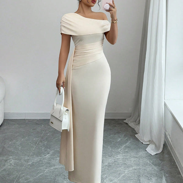 Woman in a beige off-shoulder gown holding a white handbag in a room with gray flooring and white walls.